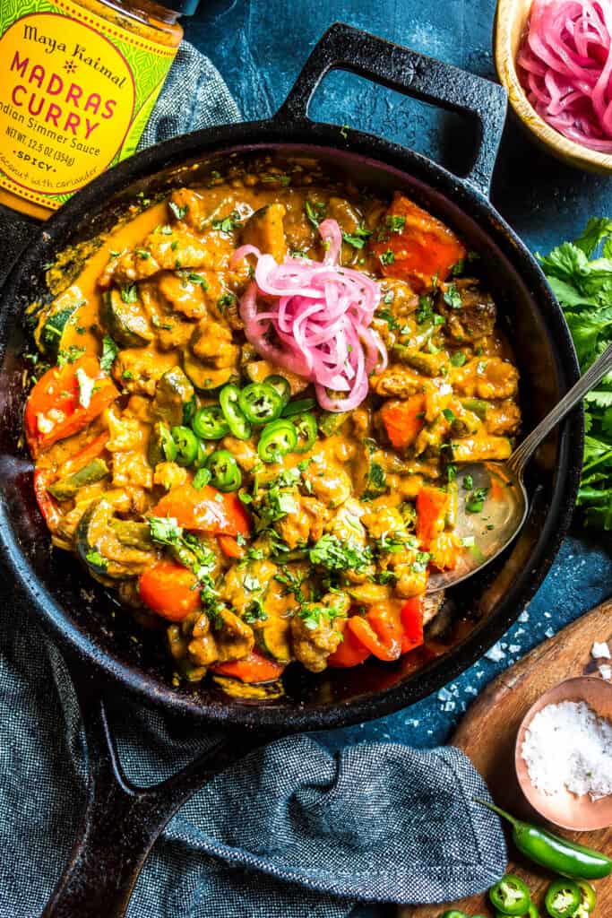 a cast iron skillet on a blue backdrop. The skillet is filled with chicken curry and topped with jalapenos, pickles onions, and cilantro. There is a jar of Maya Kaimal simmer sauce in the lefthand top corner and a variety of toppings around the right.