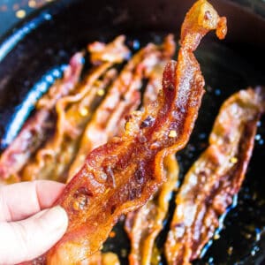 a slice of Keto Millionaire's Bacon being held above a skillet of more bacon