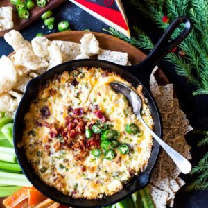 Spicy gouda bacon dip in a skillet on a platter surrounded by pork rinds and vegetable sticks.