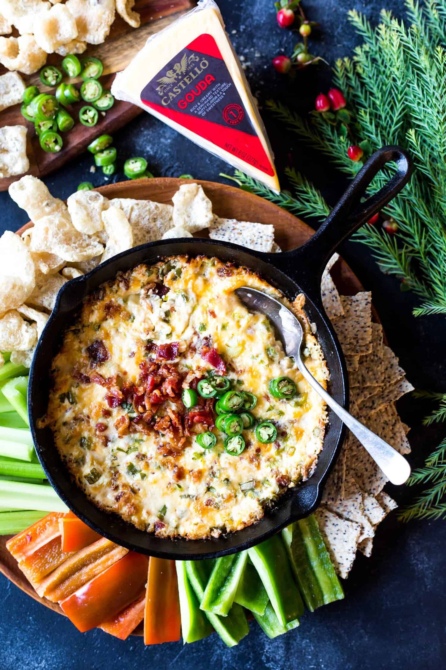 Spicy guoda bacon dip in a skillet on a platter surrounded by pork rinds and vegetable sticks.