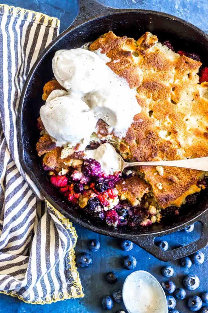 Keto Cobbler in a cast iron skillet topped with vanilla ice cream and a spoon is scooping out a bite of the berries