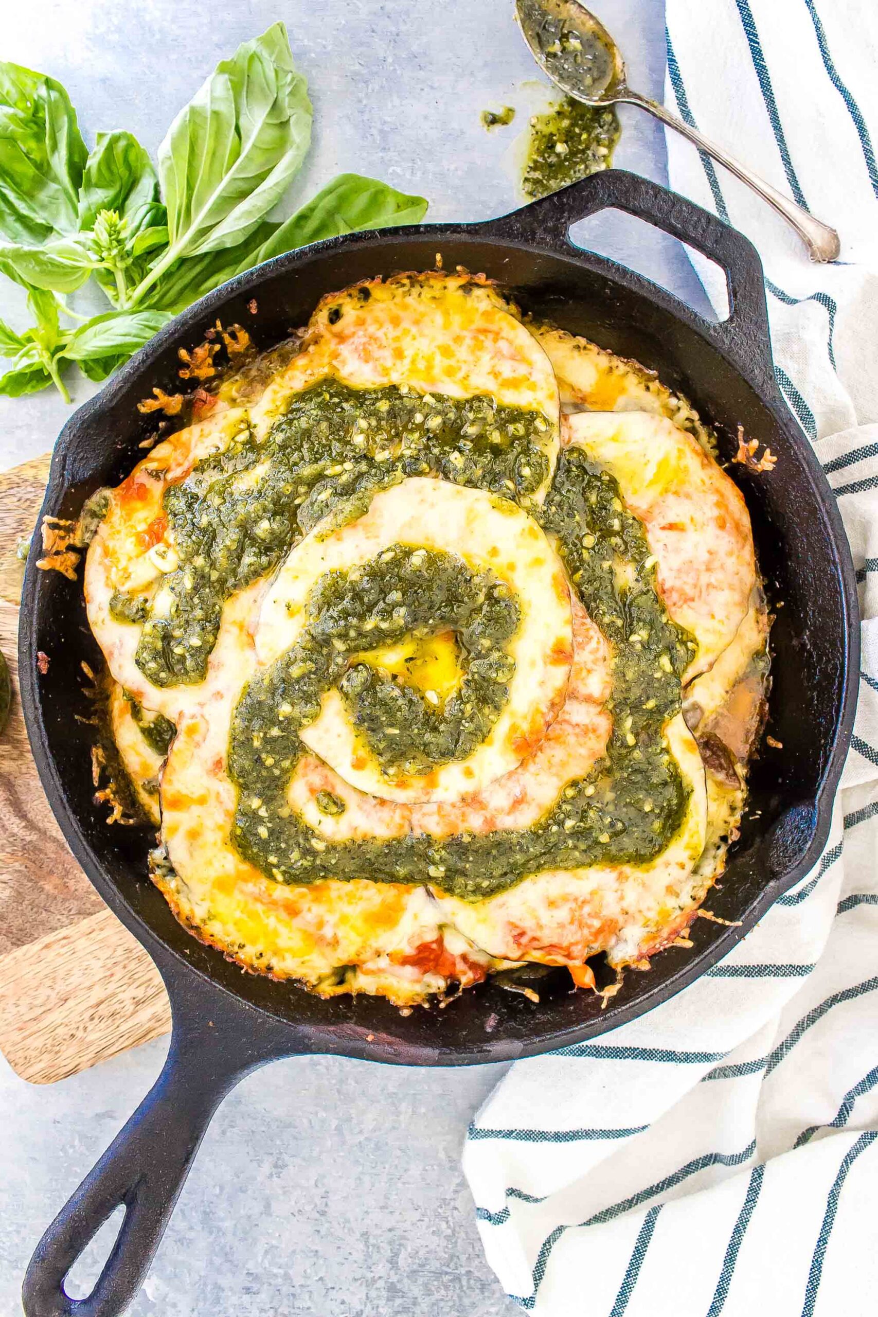 Keto Lasagna in a cast iron skillet with a pesto swirl on top, there is a striped tea towel to the left and basil leaves in the top left corner