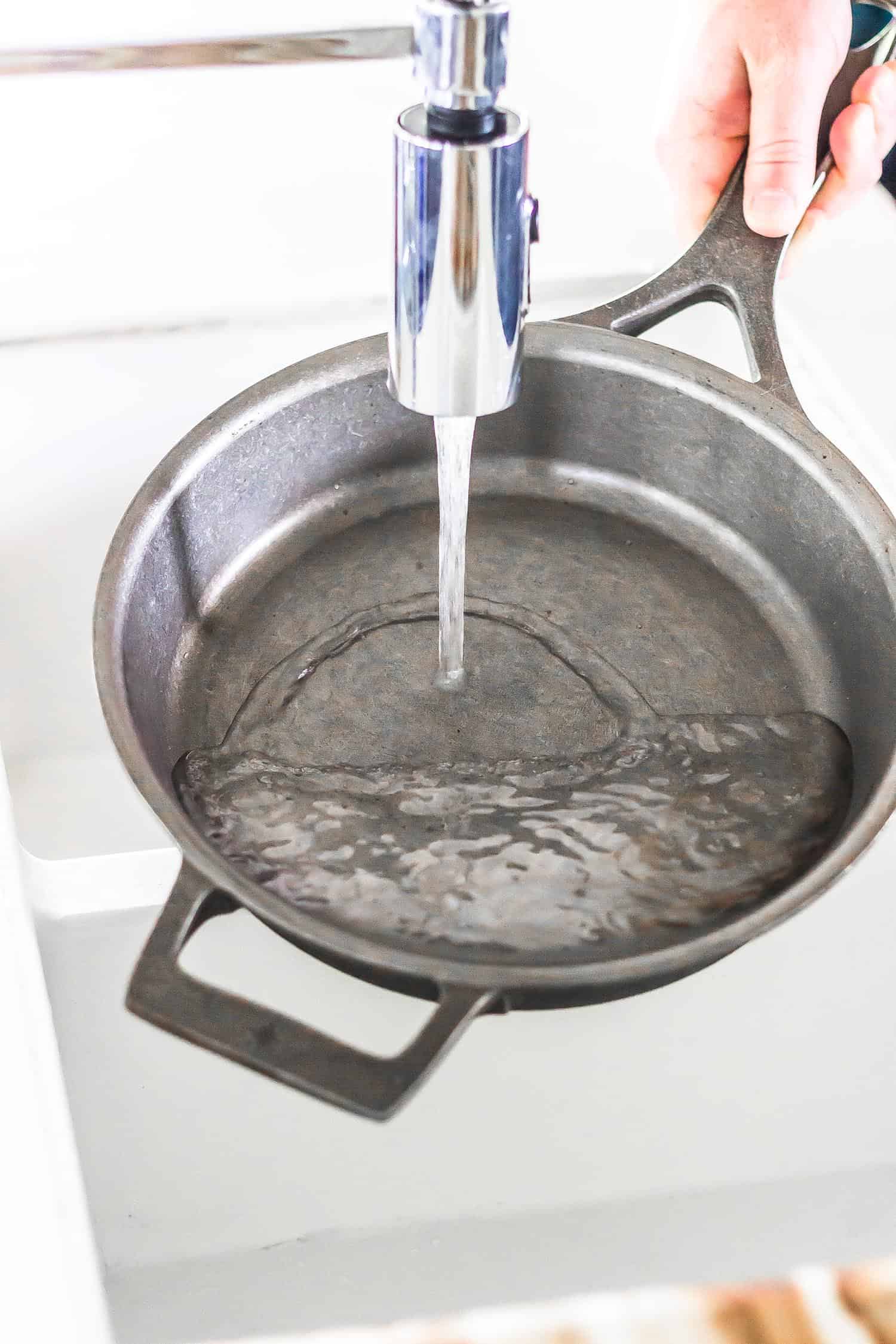 cast iron skillet being held under a kitchen faucet with water pouring into it
