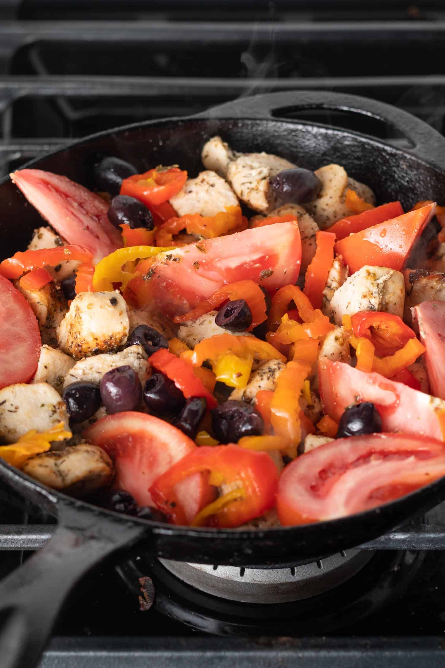 chicken, olives, tomatoes, and peppers in a cast iron skillet on top of a gas stove