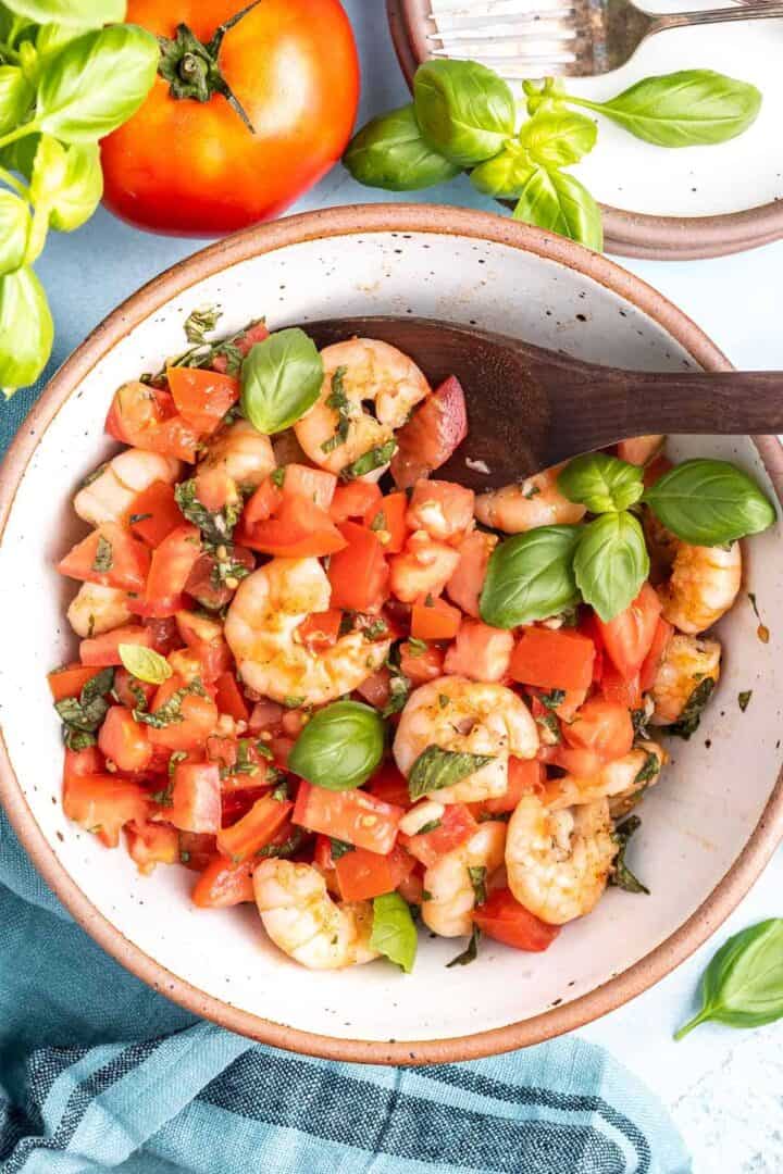 Grilled Shrimp Bruschetta in a bowl with a wooden spoon, there is a tomato in the background
