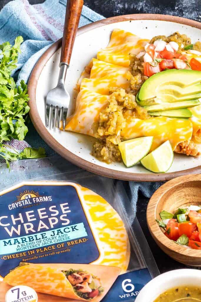 Keto Cheese Wrap Enchiladas on a plate topped with sliced avocado, pico de gallo, and limes. There is a package of Crystal Farms Cheese Wraps in the bottom lefthand corner.
