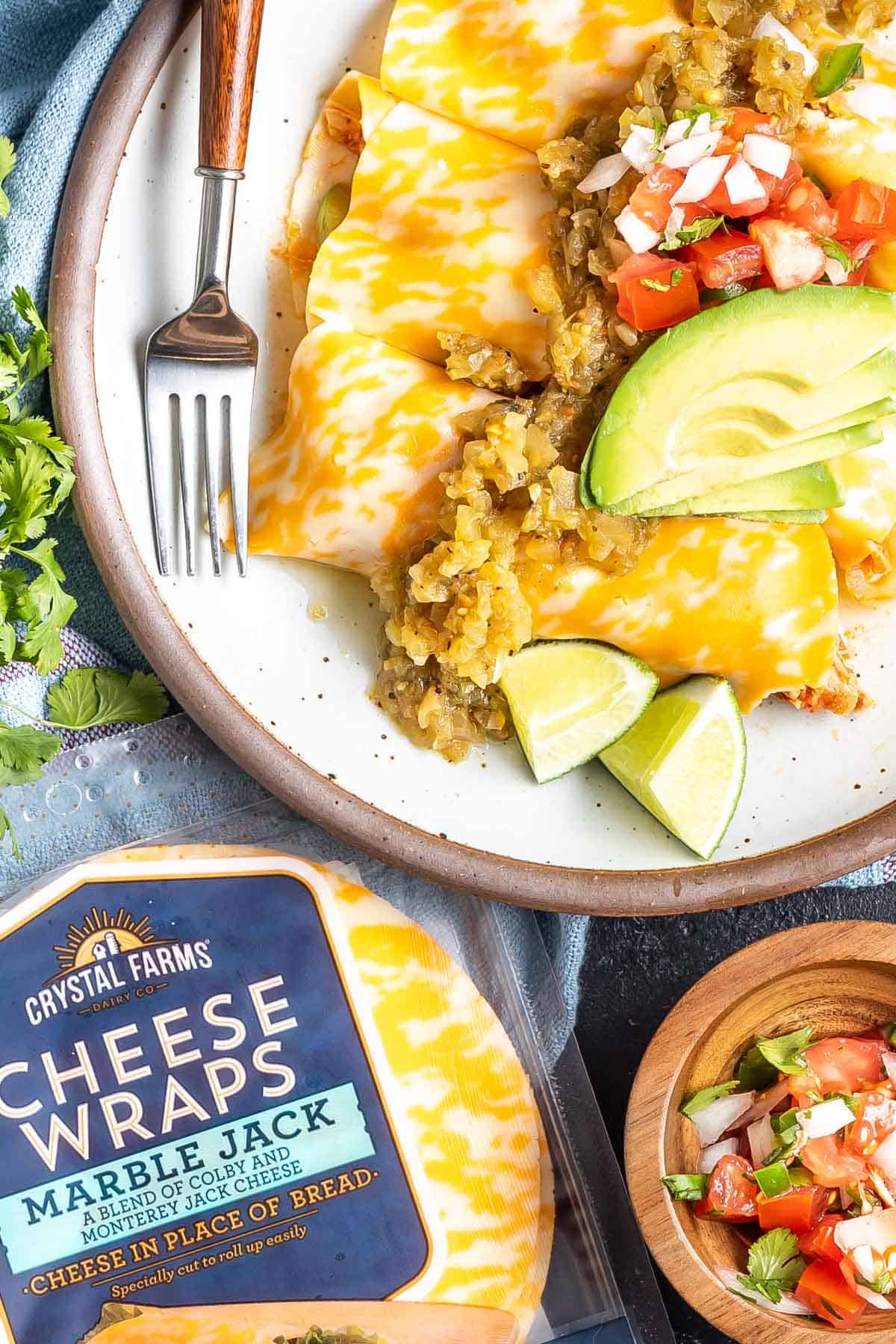 Keto Cheese Wrap Enchiladas on a plate topped with sliced avocado, pico de gallo, and limes. There is a package of Crystal Farms Cheese Wraps in the bottom lefthand corner.