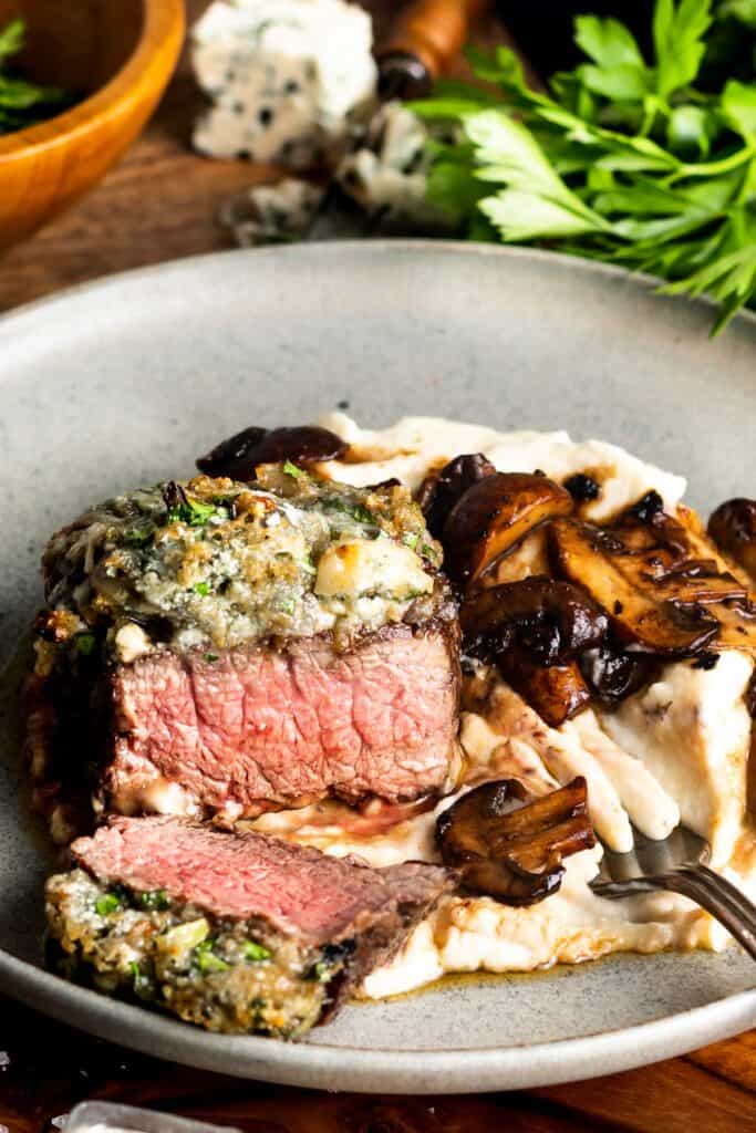 Blue Cheese Crusted Filet with Mashed Cauliflower and Balsamic Mushrooms on a grey plate on wooden saucer with a fork on the plate and parsley and crumbled blue cheese in the background