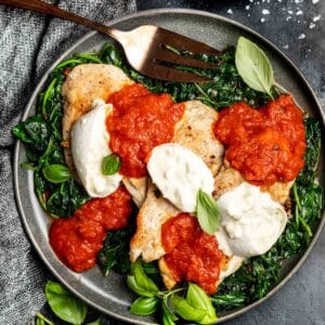 Keto Cheesy Marinara Chicken on a plate on a black background with fresh basil leaves scattered around