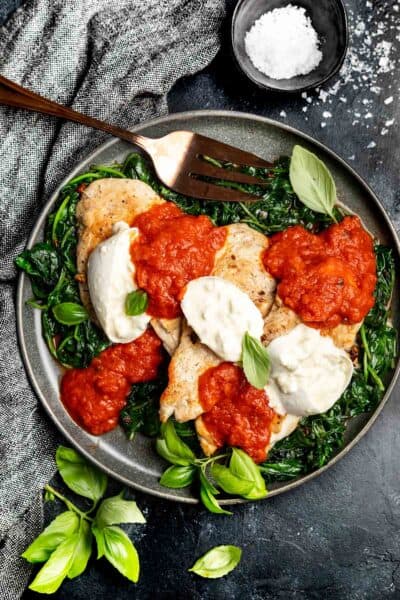 Keto Cheesy Marinara Chicken on a plate on a black background with fresh basil leaves scattered around