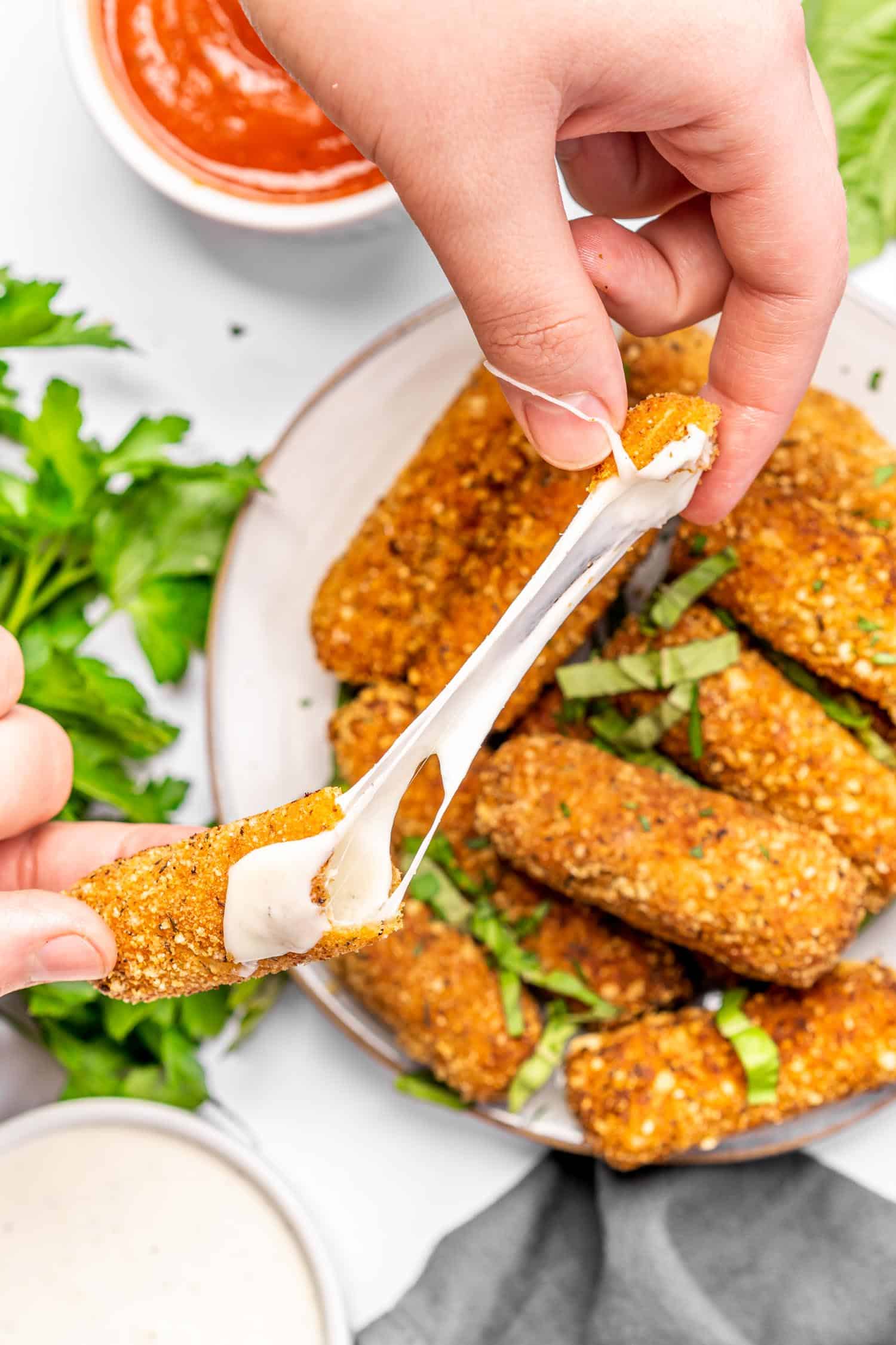 Keto Cheese Sticks being pulled apart to show a long pull of cheese in the middle. There is a plate of cheese sticks in the background along with fresh parsley and marinara dipping sauce