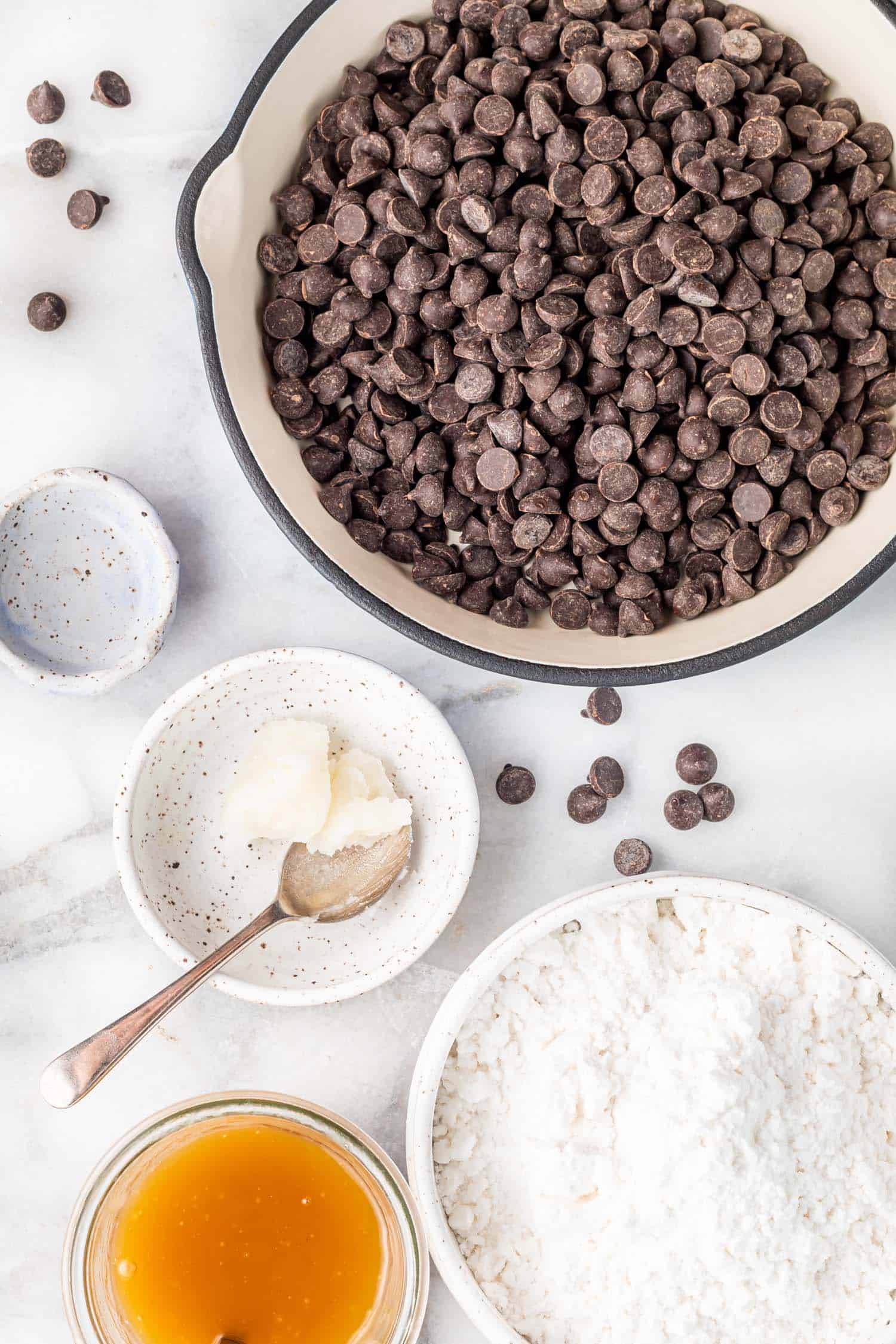 Keto Peppermint Patty ingredients - chocolate chips in a small enameled cast iron skillet, peppermint oil in a small blue bowl, coconut oil in a small white bowl, powdered sugar-free sweetener in a white bowl, and low-carb condensed milk in a jar