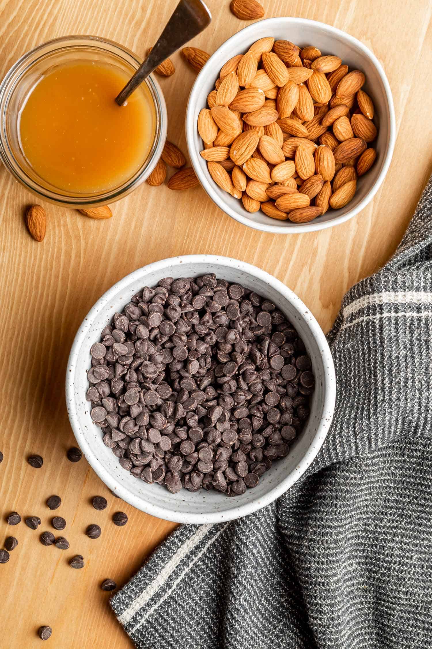 a bowl of chocolate chips, a bowl of almonds, and a jar of caramel on a wooden backdrop