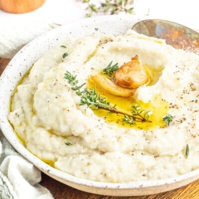 Keto Mashed Cauliflower in a white bowl topped with whole roasted garlic cloves, a sprig of Thyme, and some props to the side