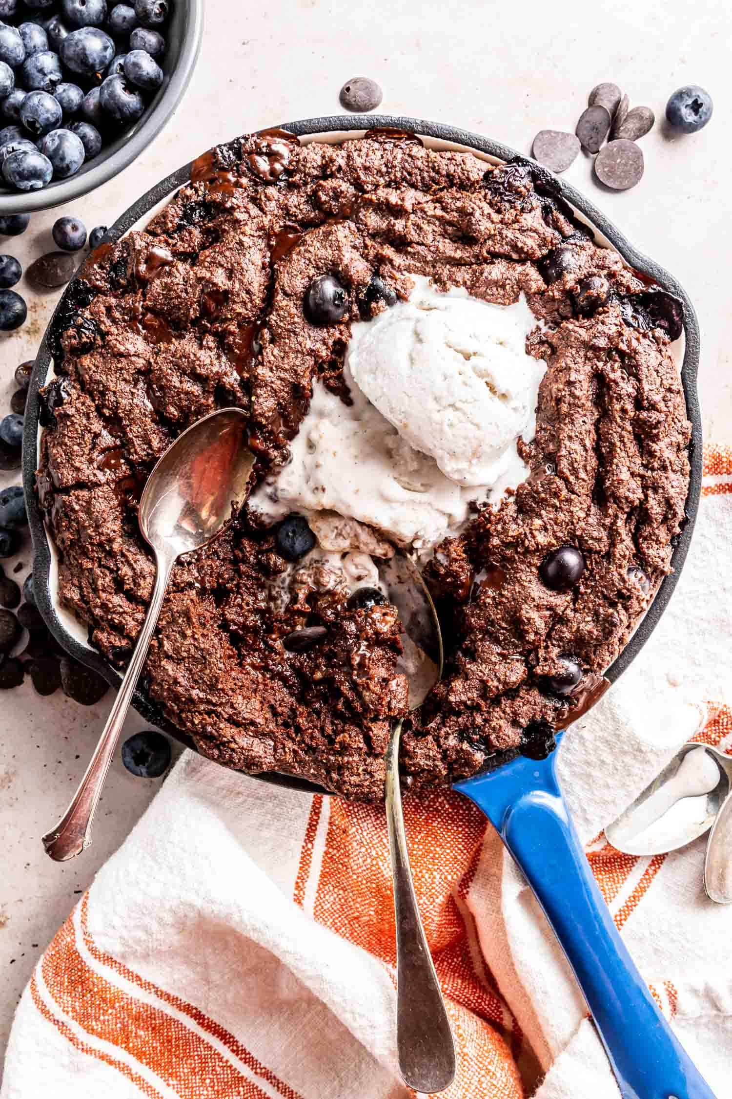 Blueberry Hot Fudge Cake in a blue enameled cast iron skillet topped with ice cream with two spoon dug into the center.