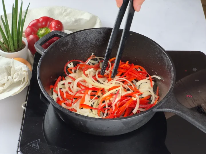 Thinly sliced yellow onions and red peppers getting stirred in a cooking pan.