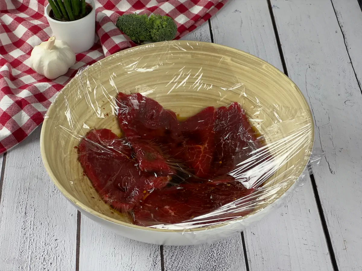 Beef pieces left for marinating in a large bowl covered by plastic foil.