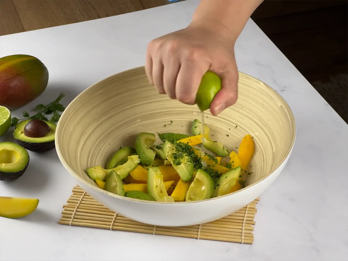 Lime being squeezed in a bowl containing sliced mangoes, avocados, and finely chopped cilantro.