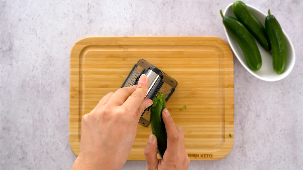 A step-by-step guide to preparing homemade keto tzatziki: grating fresh cucumbers on a cutting board.