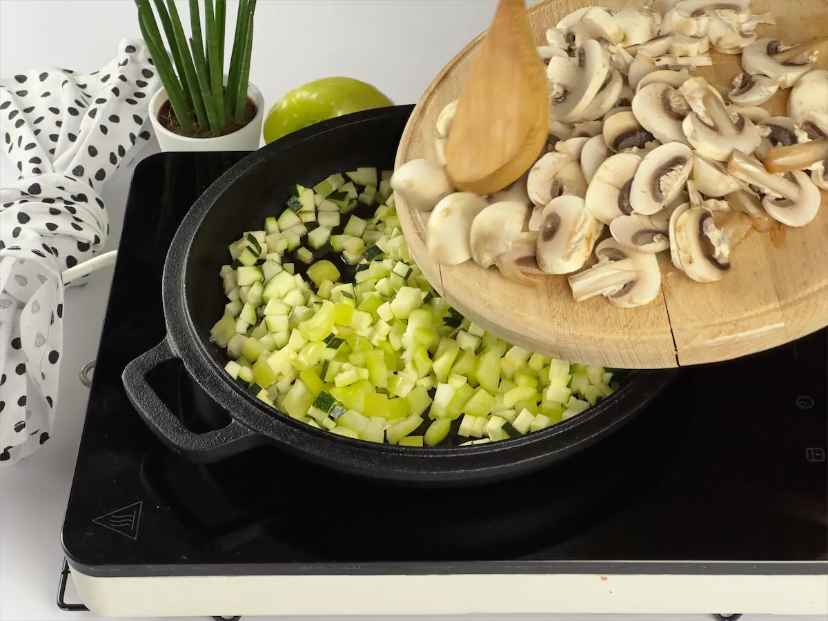Mushrooms being added in a cast iron pan with finely chopped bell peppers and zucchini.