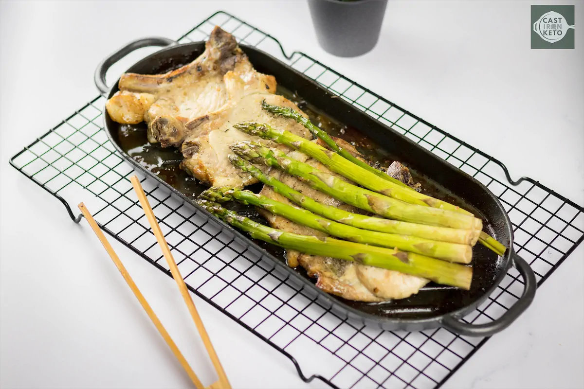 Aesthetic arrangement of keto baked pork chops with asparagus on a cast iron baking dish on kitchen table.