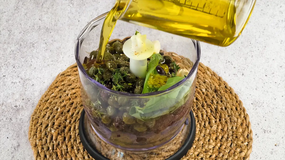 Adding olive oil to the mixture of ingredients to make Classic Olive Tapenade.