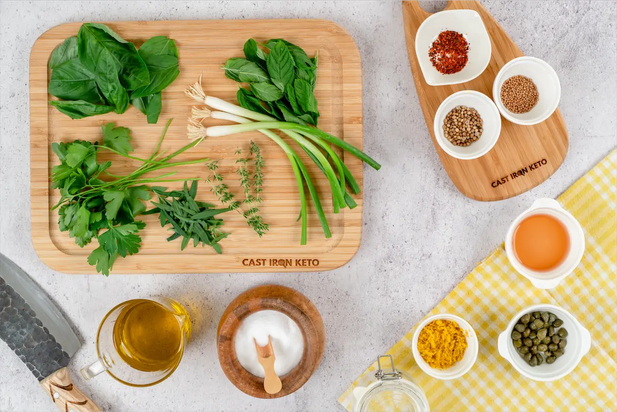 Ingredients like fresh parsley, mint, spring onion, basil, fresh thyme, tarragon, mustard seeds, coriander seeds, chilli flakes, vinegar placed on a table.