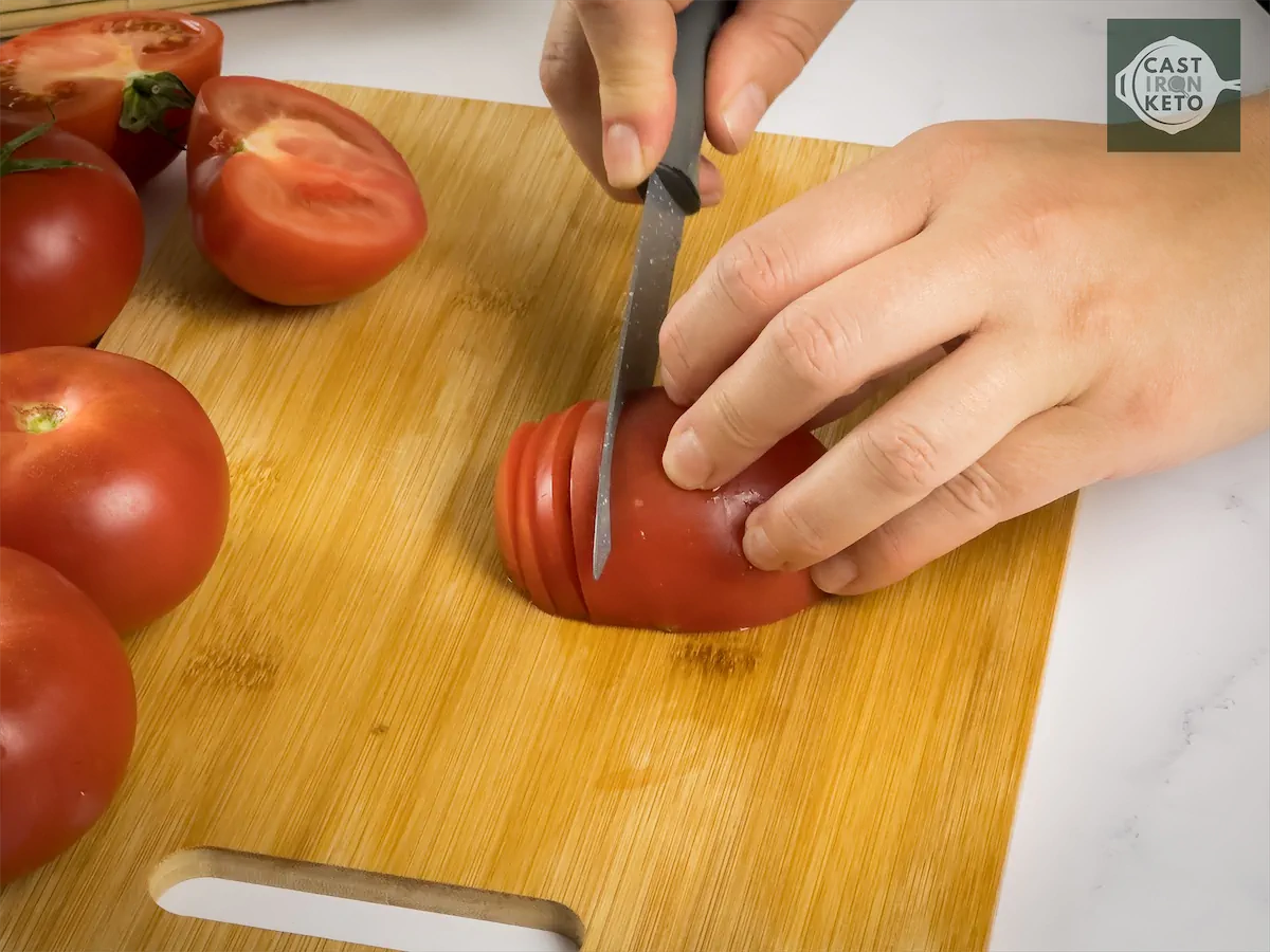 Tomato being thinly sliced by a knife on chopping board.