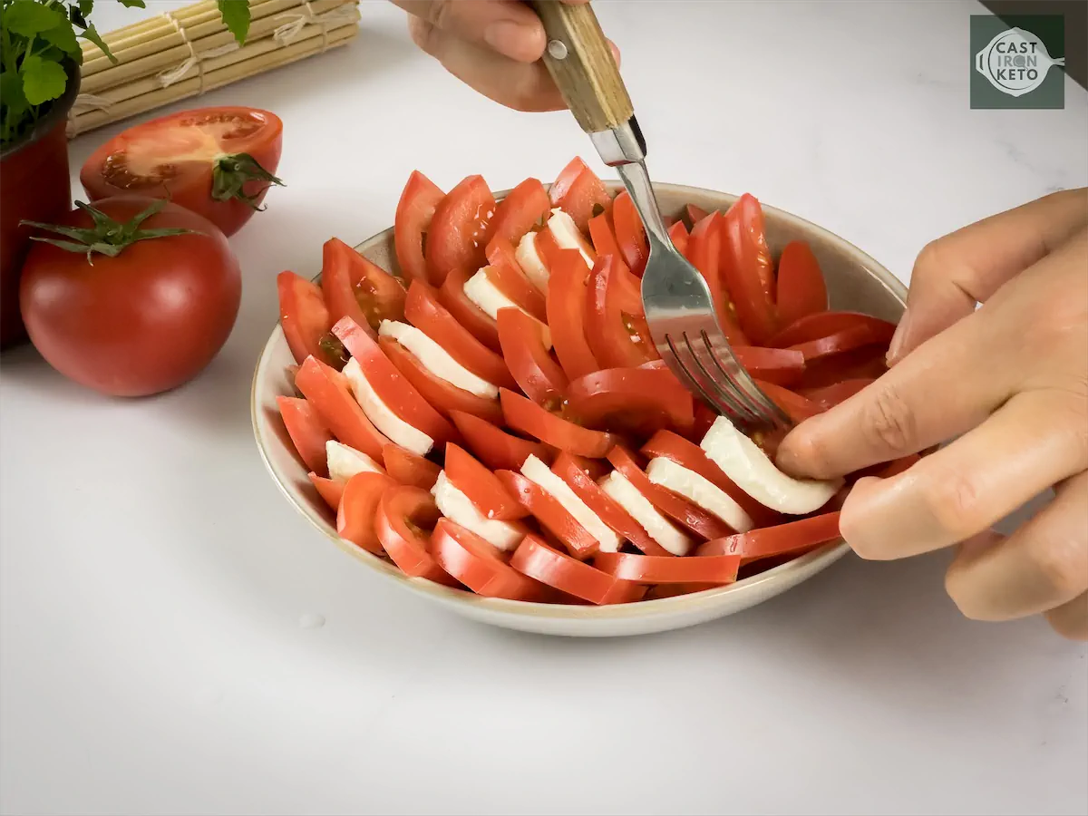 Placing slices of mozzarella in between sliced tomatoes placed in a bowl.