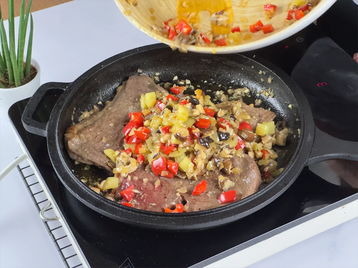 Veggies like peppers, onions and mushrooms being added in a cast iron pan with steaks.