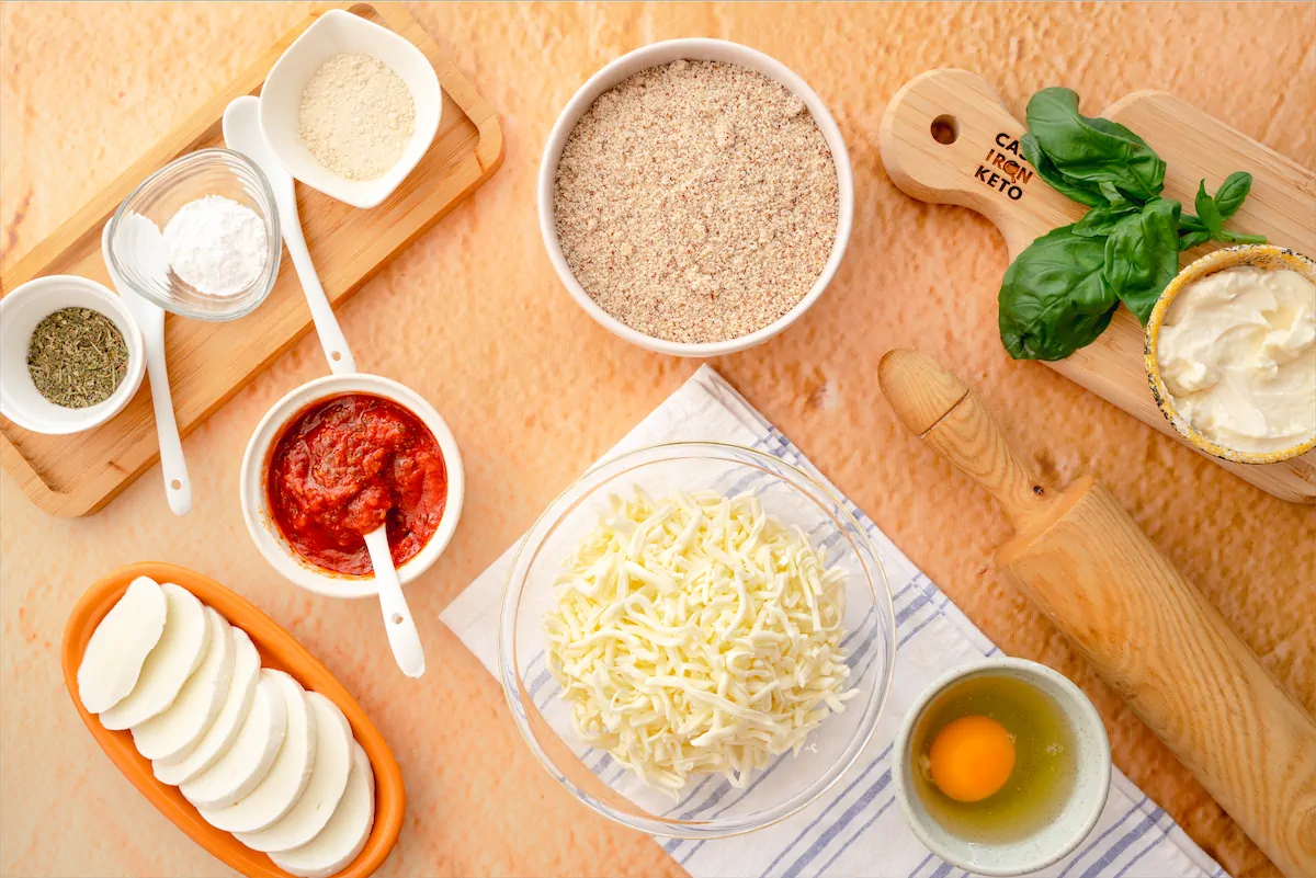 Ingredients like grated mozzarella cheese and slices, cream cheese, almond meal, egg, tomato sauce, basil leaves made ready on the table to prepare keto pizza.