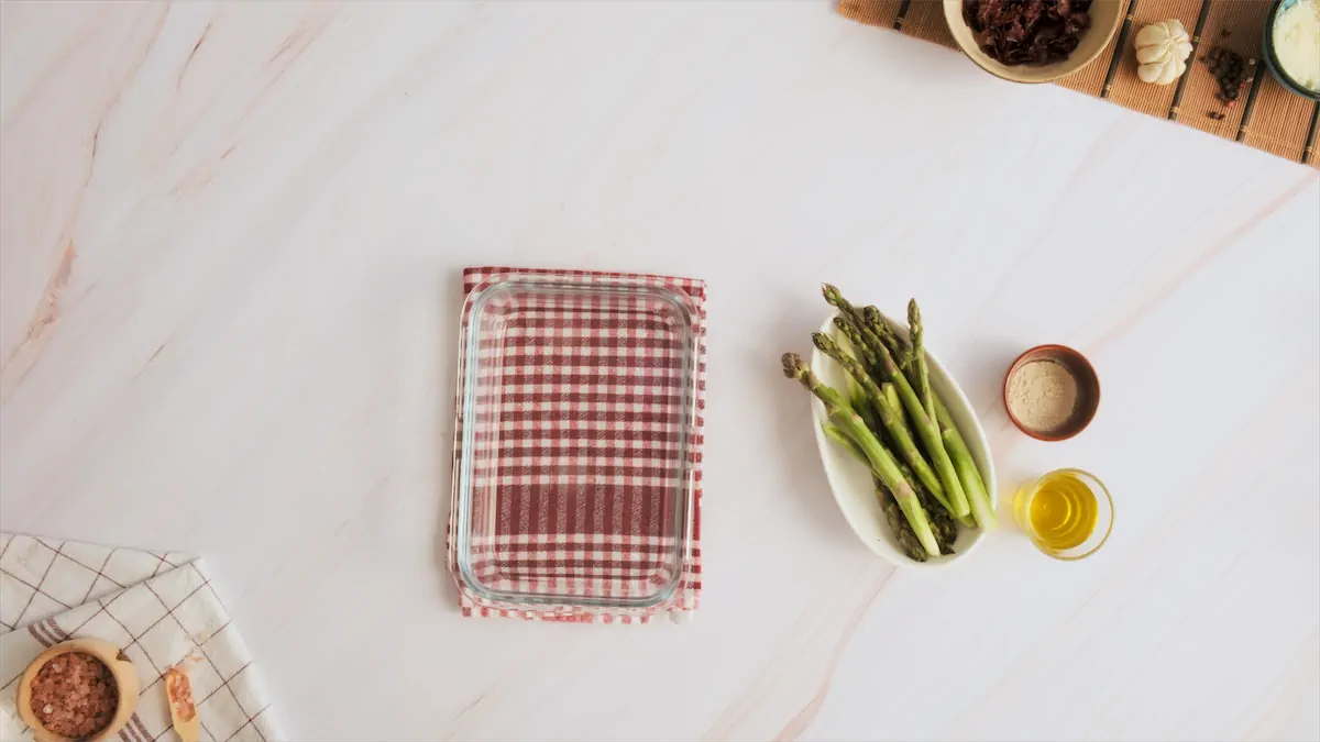 Glass baking dish next to ingredients like asparagus, olive oil, and garlic powder on kitchen table.