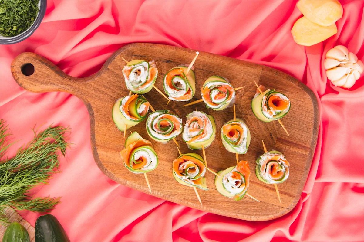 Overhead shot of keto smoked salmon cucumber rolls placed on a wooden serving tray accompanied by some vegetables on kitchen table.