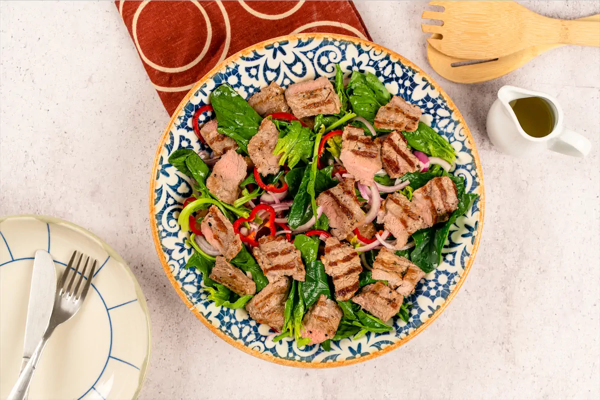Steak spinach salad on kitchen table next to cutleries.