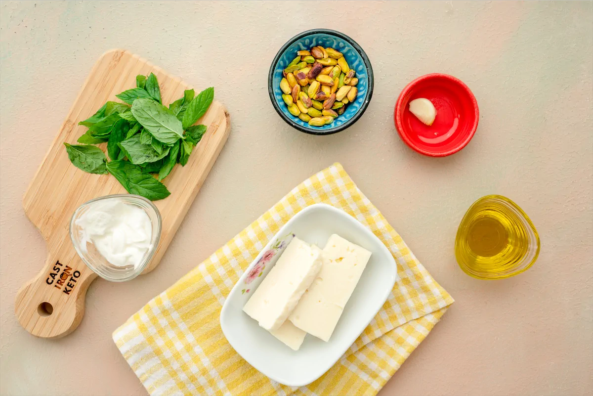 Pistachios, fresh mint leaves, feta cheese, olive oil, garlic clove, and Greek yogurt made ready on the table to prepare Tirokafteri recipe.