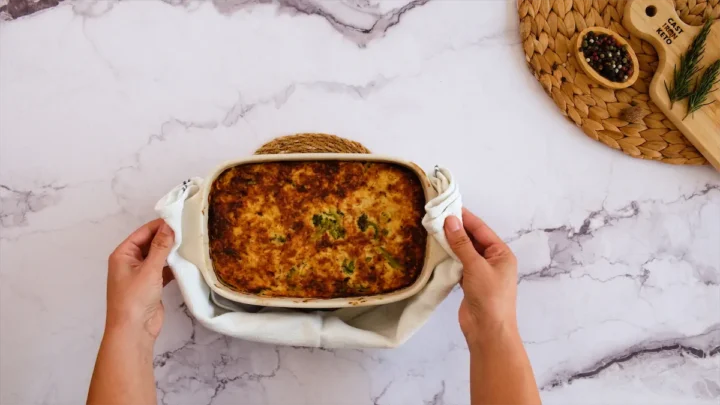 Baked casserole recipe in a baking dish safely caught with two hands using a cloth.