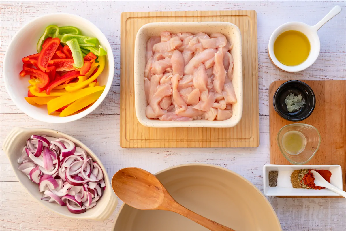 Sliced chicken breasts, sliced bell peppers, sliced onions, olive oil, lime juice, minced garlic, chili powder, oregano, cumin, salt, and black pepper spread out on kitchen table.