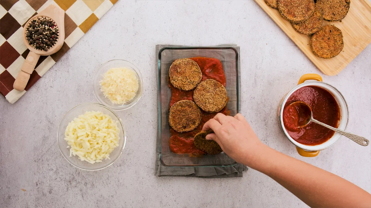 Placing the baked eggplant slices in a baking dish containing tomato sauce.