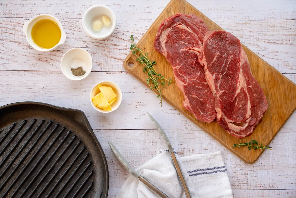 Sirloin steaks, olive oil, garlic cloves, fresh rosemary, fresh thyme, salt, black pepper, and butter spread out on kitchen table for the preparation of sirloin steak recipe.