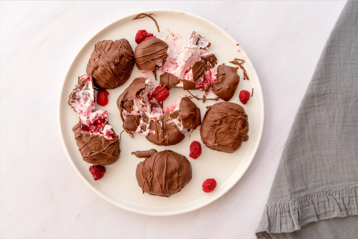 Chocolate-covered frozen yogurt bites with some raspberries are arranged on a plate and some of them cut open.