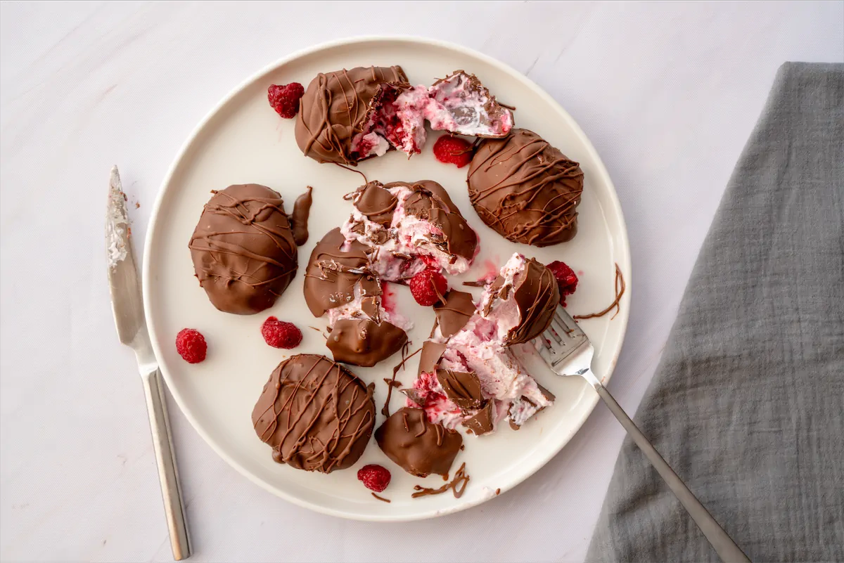 Dark-chocolate-covered frozen yogurt bites and raspberries on a plate where some are cut with a fork and a knife.