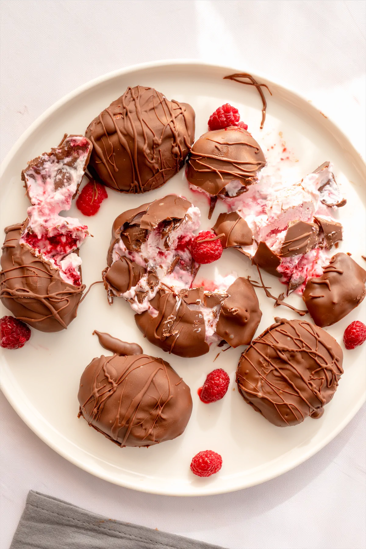 Dark-chocolate-covered frozen yogurt bites decorated with some raspberries are on a plate, some of them sliced open.
