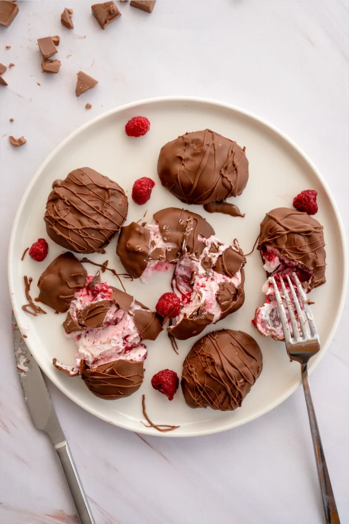 Frozen yogurt bites covered in melted chocolate are on a plate, some sliced open to reveal raspberries inside.