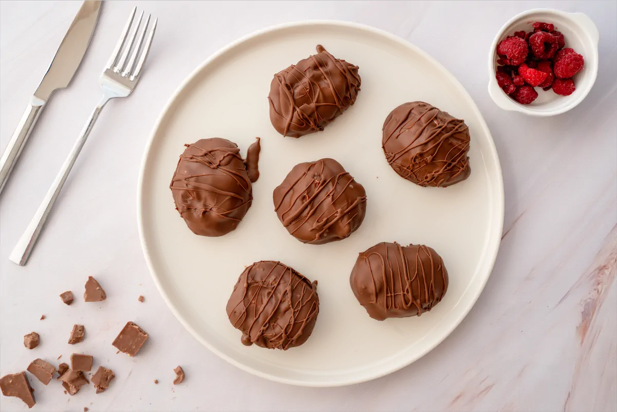 Chocolate-covered frozen yogurt bites are arranged on a plate besides a bowl of some raspberries and cutlery.