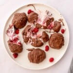 A plate of chocolate-covered frozen yogurt bites decorated with raspberries, some are smashed open.