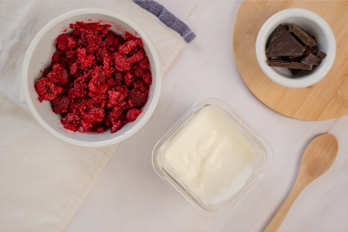 A bowl of yogurt, chopped raspberries, dark chocolate and a wooden spoon on a table.