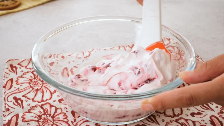 In a bowl, yogurt and raspberries are getting mixed with a spatula.