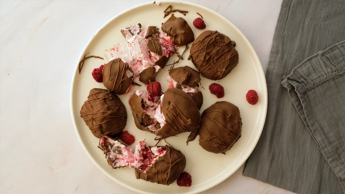 A plate of chocolate-coated frozen raspberries bites, some are smashed showing the berries inside.