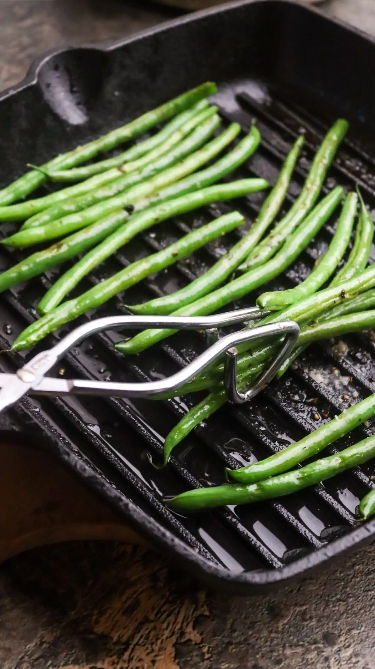 Green beans are getting turned using tongs on a cast iron grill skillet.