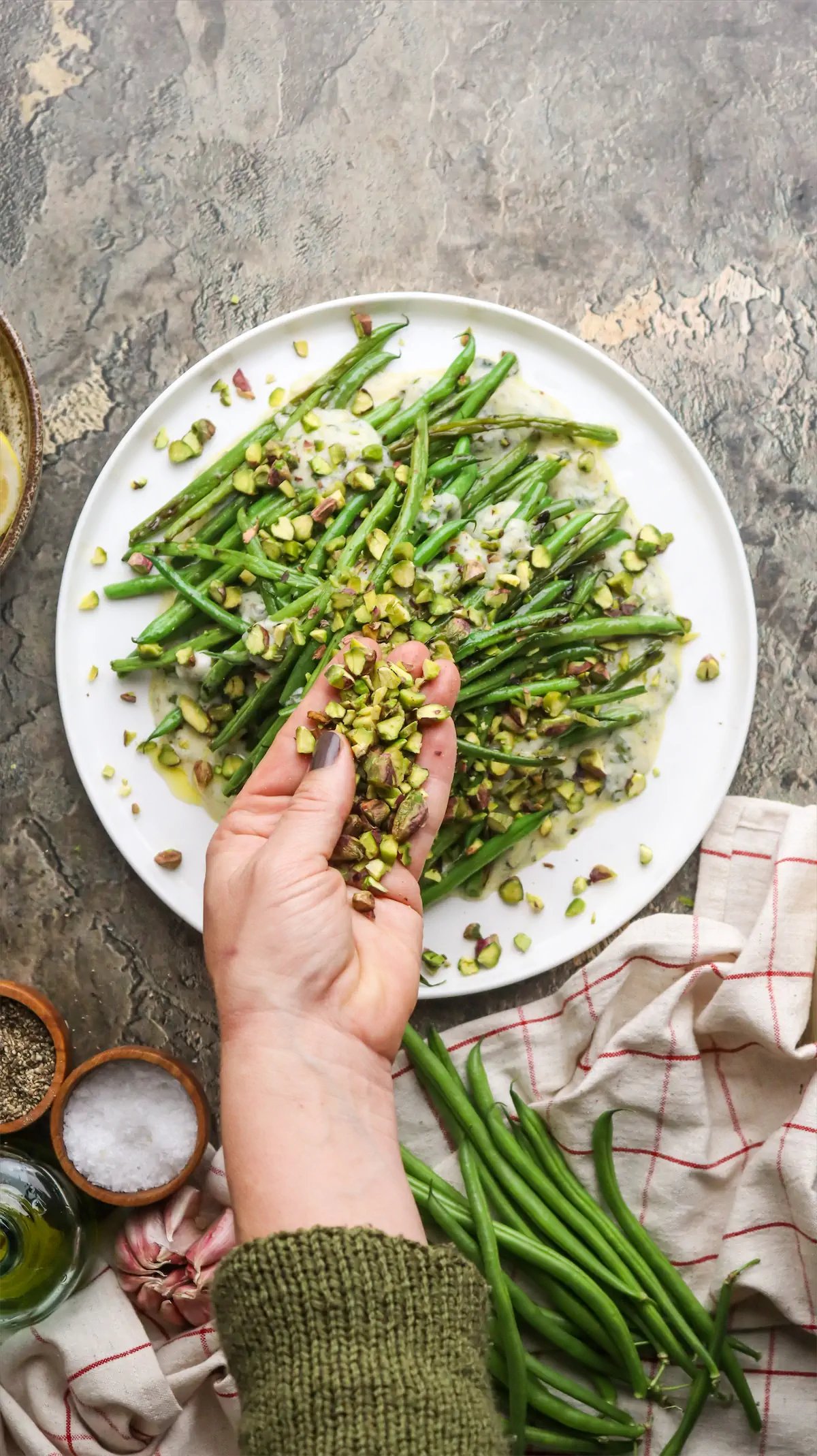 A hand sprinkling crushed pistachio over the plate of green beans with yogurt and mint dressing.